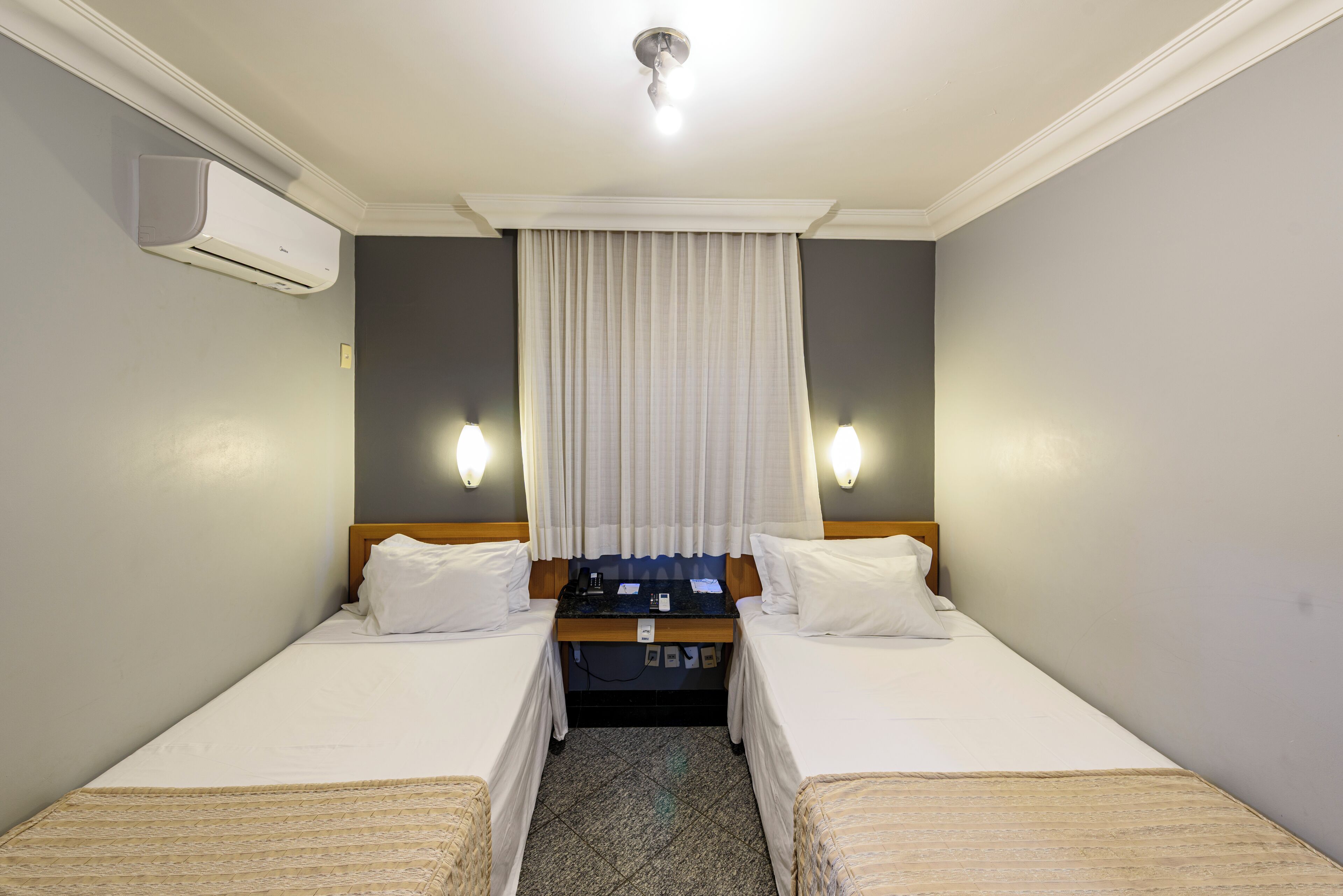 A hotel room with two single beds, a small desk with a chair, a nightstand, and a window with curtains.