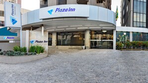 Exterior del Hotel Plaza Inn Executive, con un diseño moderno, que presenta una entrada curva, un jardín ajardinado con flores rojas y un camino empedrado que conduce a la entrada.