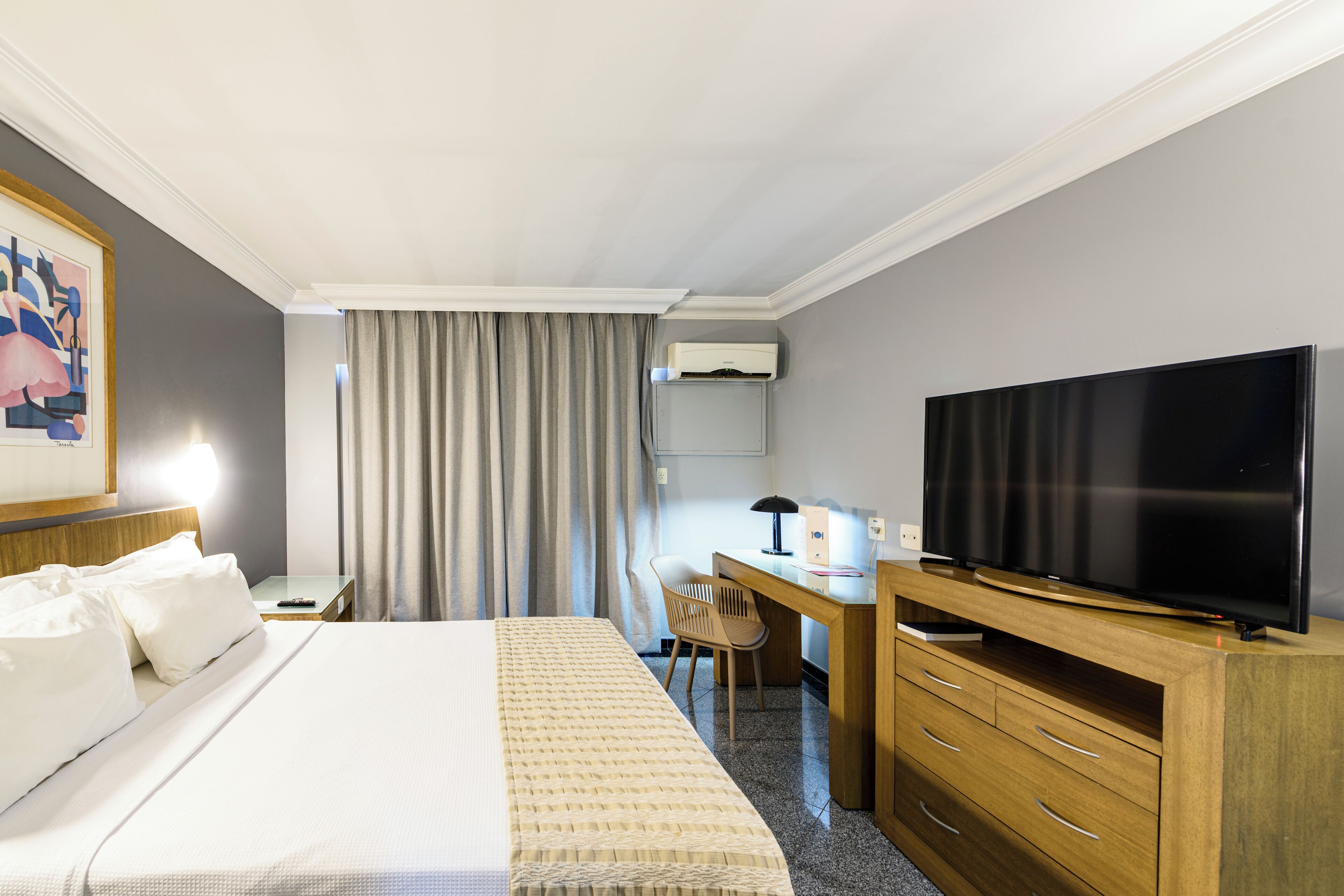 Una habitación de hotel con cama, escritorio, silla, televisor y aire acondicionado.