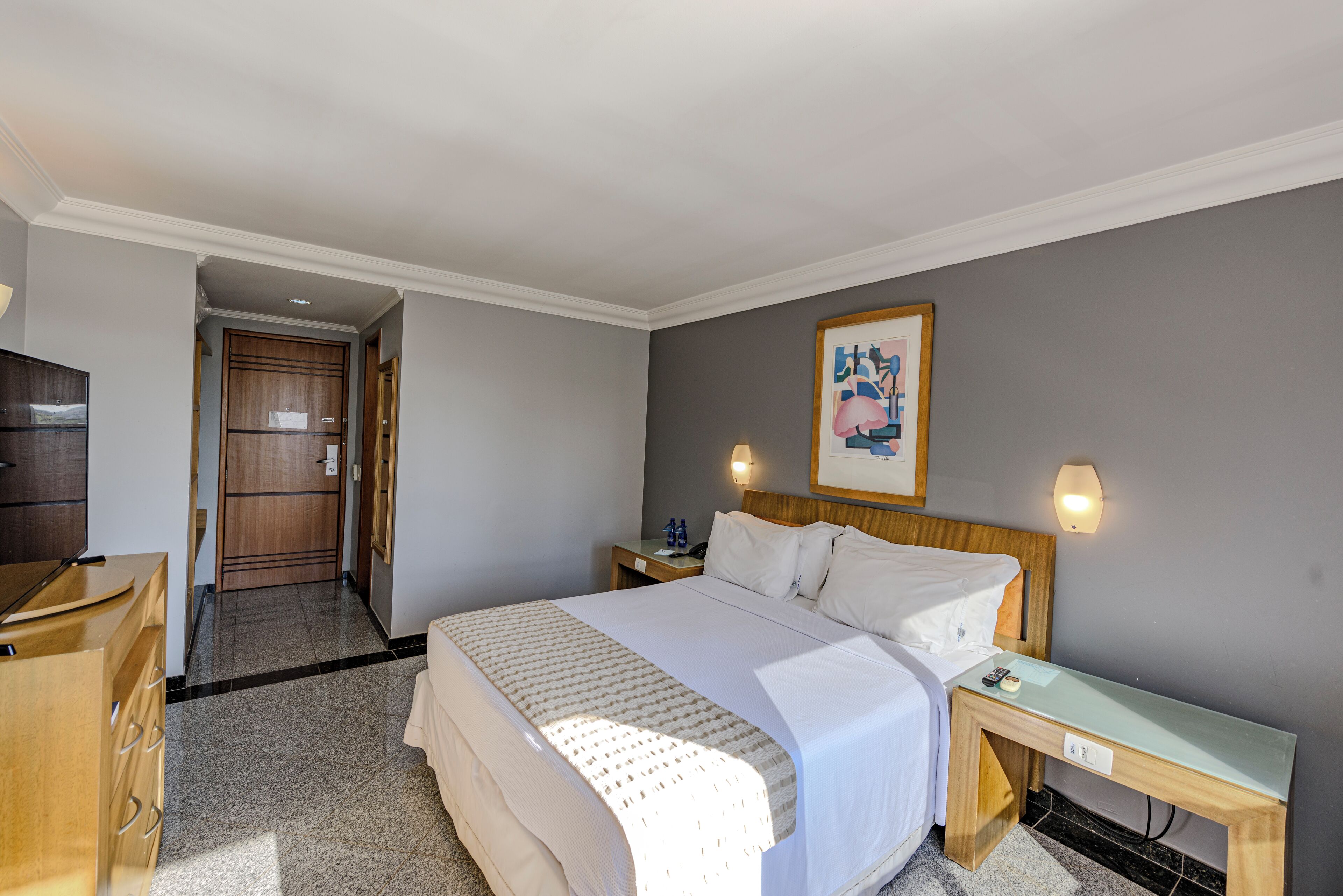A hotel room with a bed, bedside tables, a desk, and a painting on the wall.