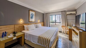 Habitación de hotel con una cama grande, un escritorio, una silla, un aparador, una mesita de noche y una ventana con cortinas.