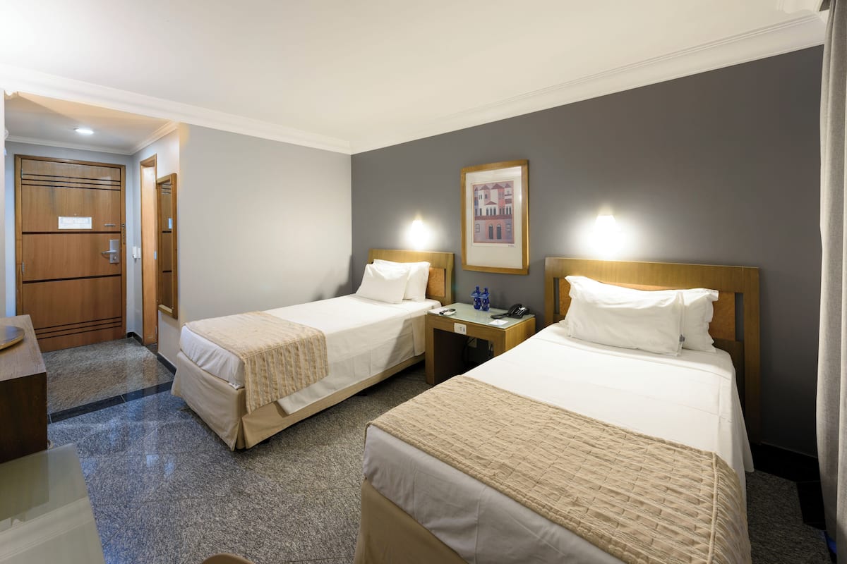 Ein Hotelzimmer mit zwei Einzelbetten, einem Holzschrank, einem kleinen Tisch mit Telefon und Lampe sowie einem gerahmten Bild an der Wand.