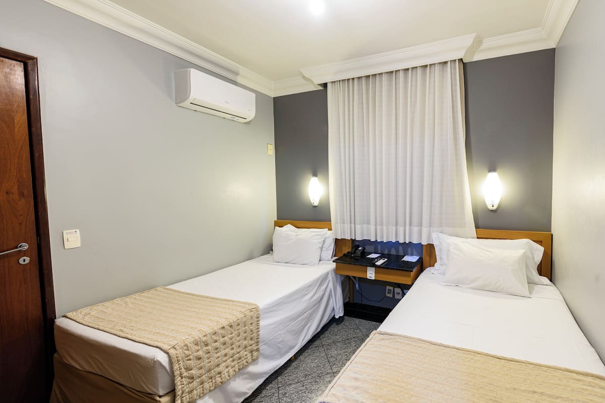 Ein Hotelzimmer mit zwei Einzelbetten, einer Holztür, einer Klimaanlage, einem Nachttisch und einem Fenster mit Vorhängen.