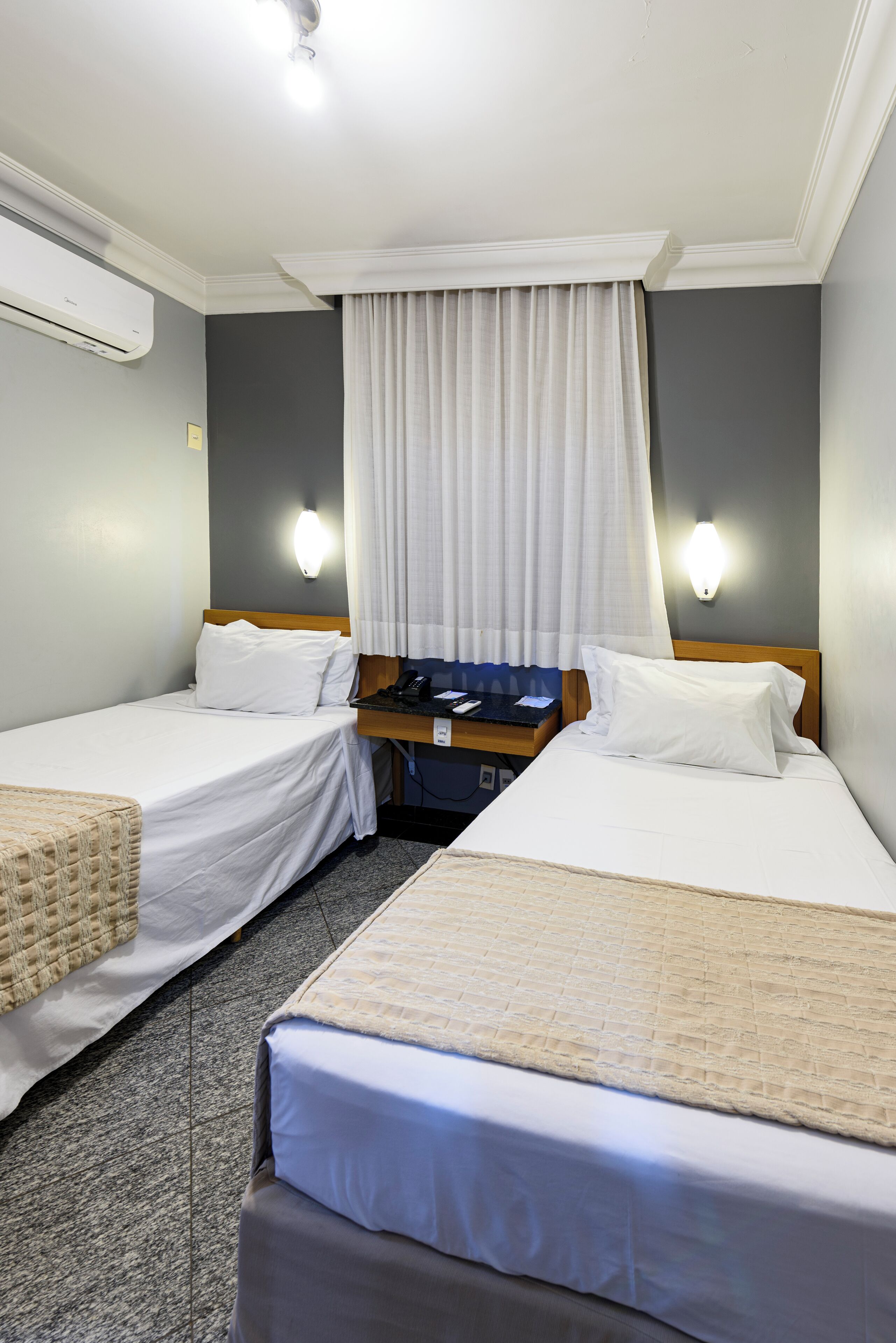 A hotel room with two beds, a nightstand, an air conditioning unit, and a window with curtains.