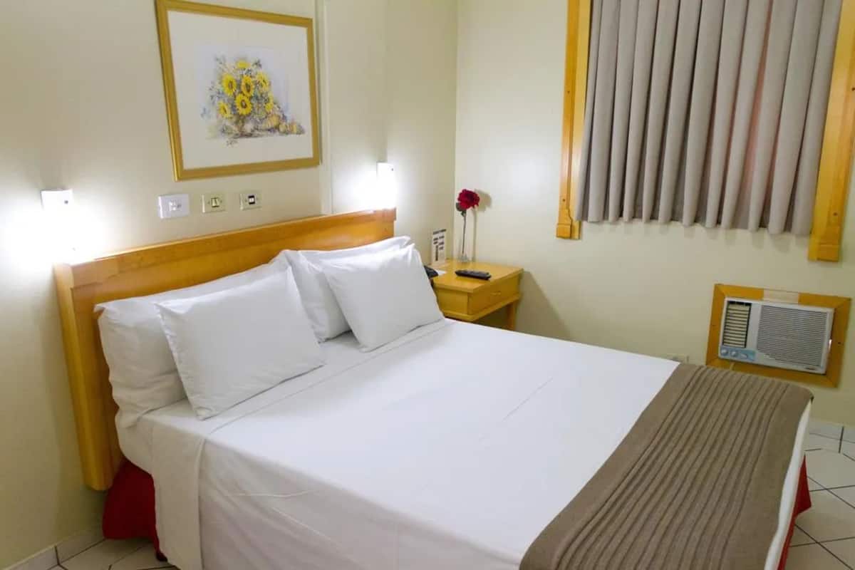 A hotel room with a wooden headboard, white bedding, a bedside table with a lamp, a framed floral artwork, a red flower vase, a window with blinds, and a wall-mounted air conditioning unit.
