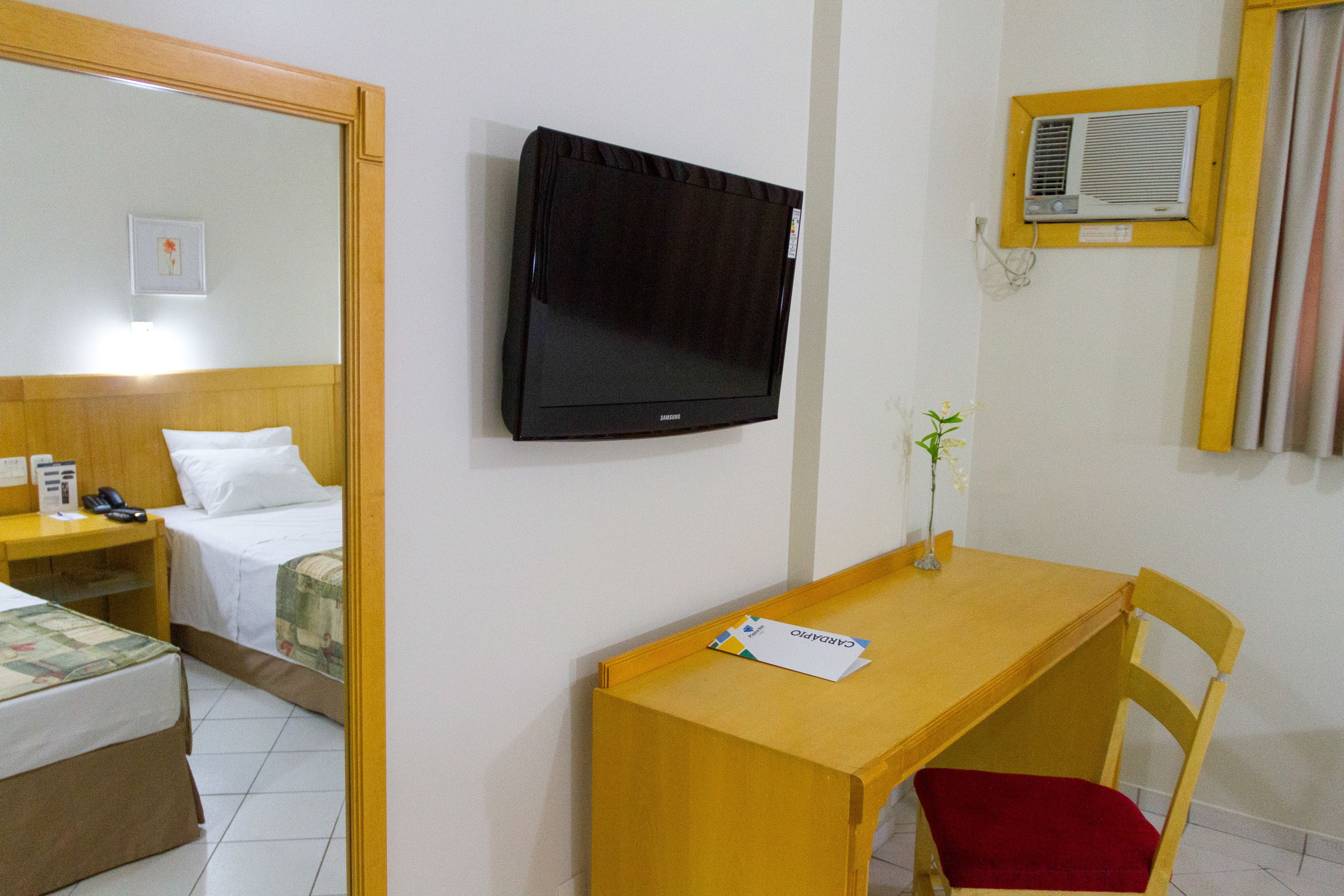 Quarto de hotel com cama, escrivaninha, cadeira, televisão e espelho.