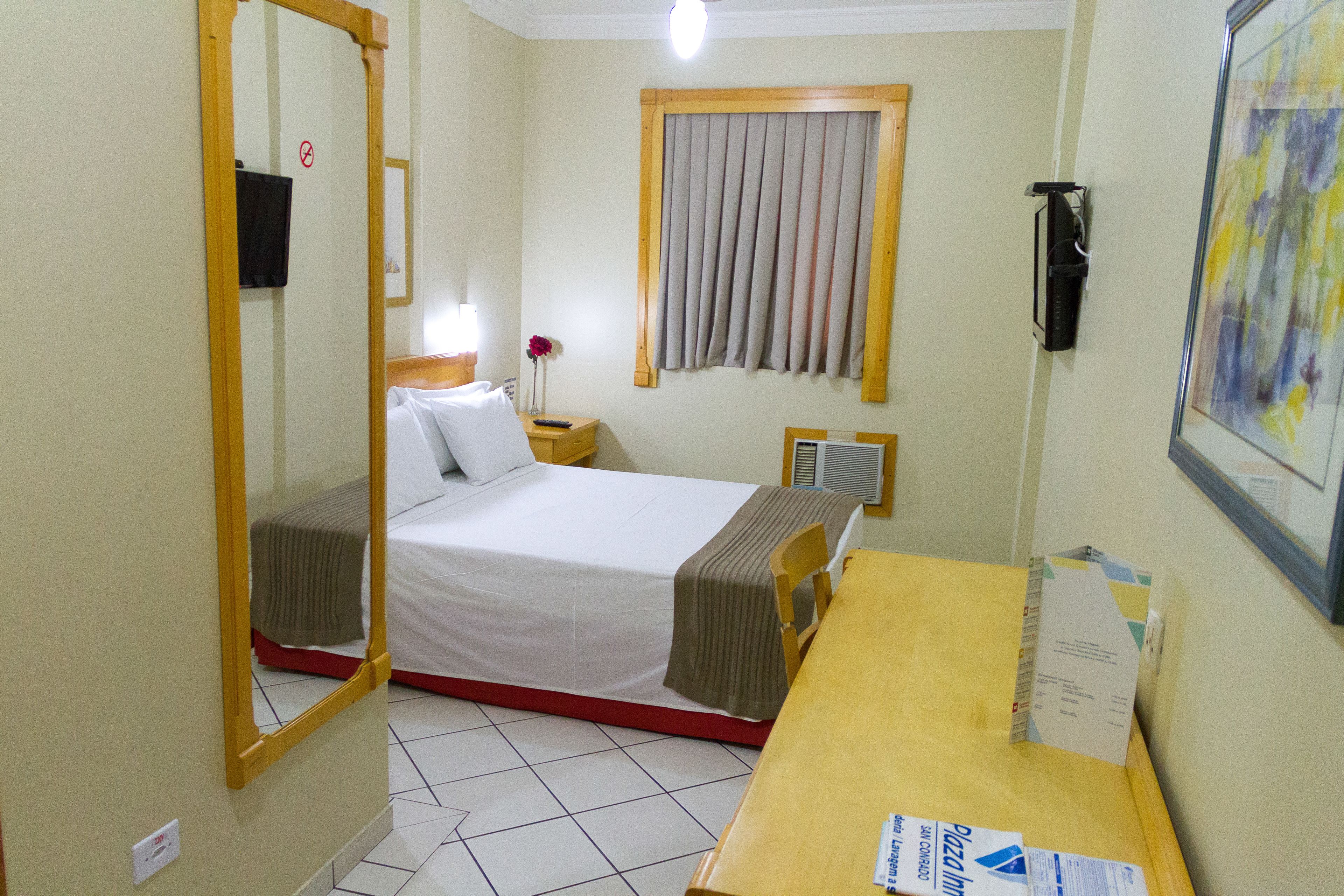 Una habitación de hotel con una cama, una mesita de noche de madera, un espejo, una ventana con cortinas, un equipo de aire acondicionado instalado en la pared y un cuadro enmarcado.
