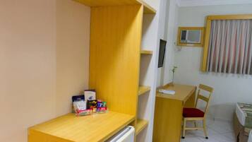 A compact room with a wooden desk, a chair, a small refrigerator, and a window with curtains.