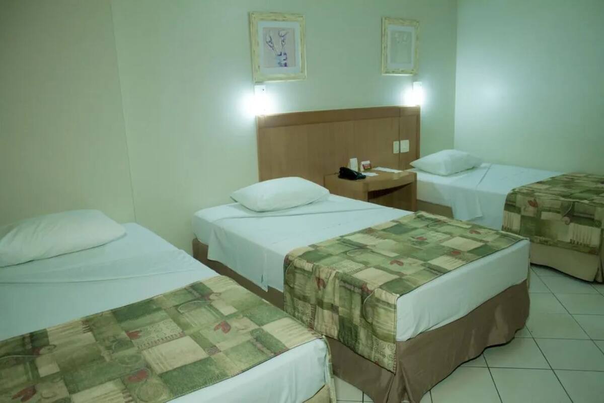 A hotel room with three single beds, each with a different style of bedding. There are bedside tables with lamps and a wall-mounted light fixture.
