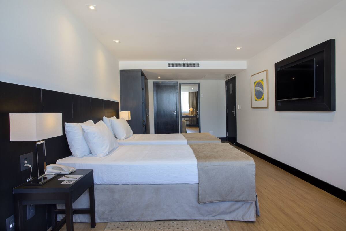 Una habitación de hotel con dos camas, un escritorio con teléfono y lámpara, un televisor montado en la pared y una puerta que da a otra habitación.