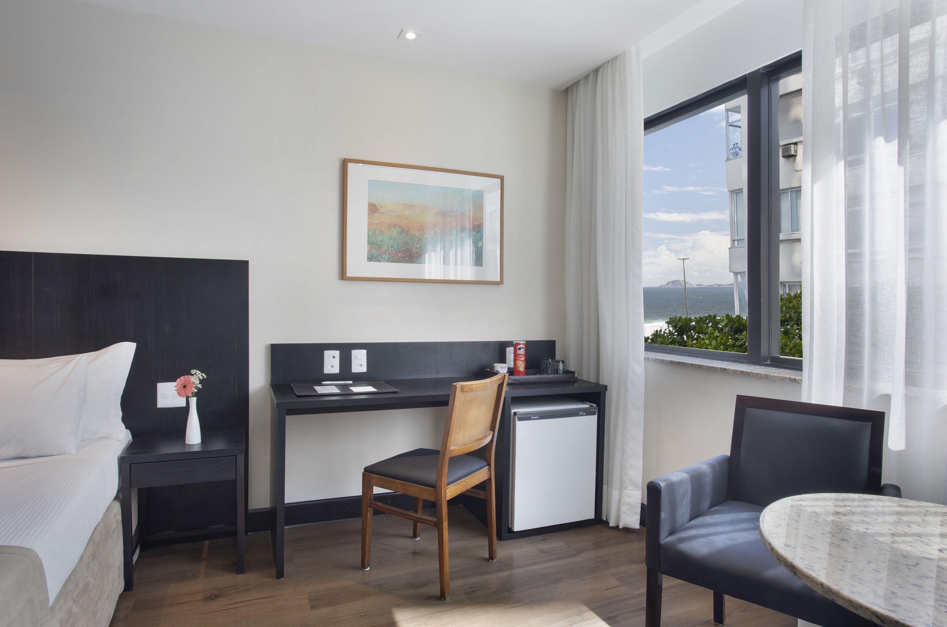 Quarto de hotel com cama, escrivaninha, cadeira, mini-frigorífico, mesa e janela com vista.