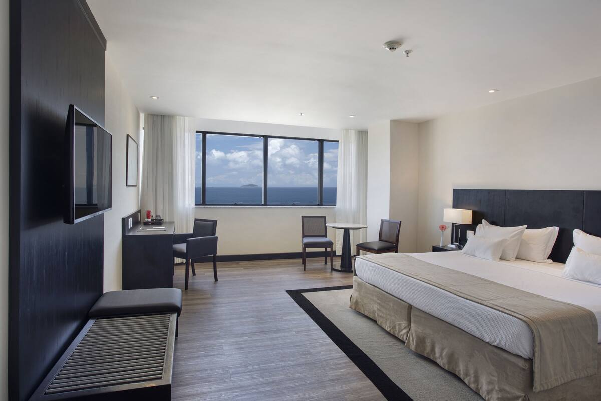 Un hotel moderno con una cama grande, un escritorio, dos sillas, una mesita pequeña y un amplio ventanal con vistas al mar.