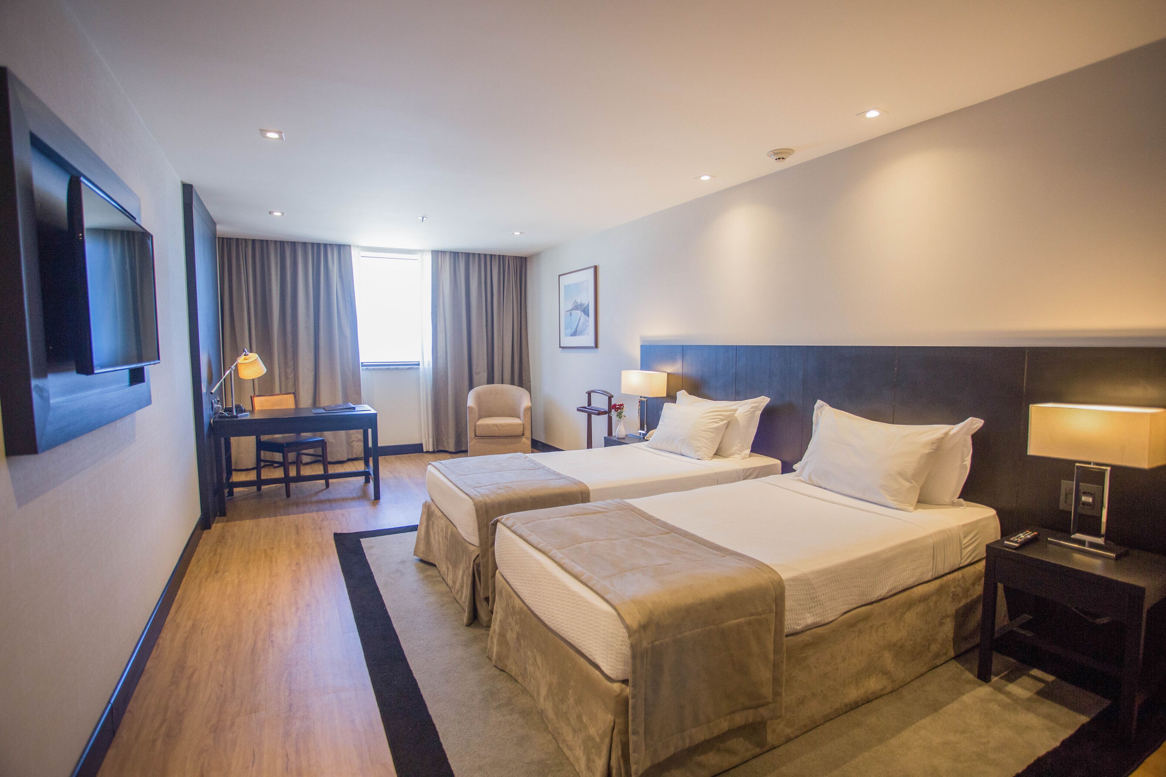 A hotel room with two beds, a desk, a chair, a TV, and a window with curtains.