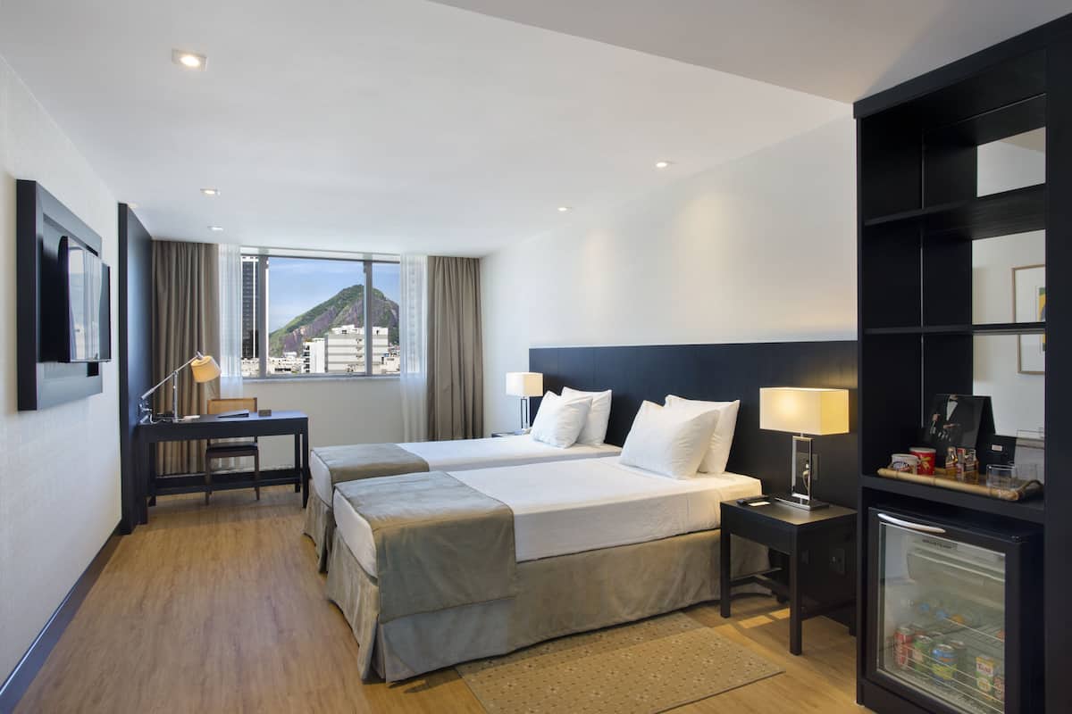 Habitación de hotel con dos camas, un escritorio, un refrigerador y un amplio ventanal con vista a la ciudad.