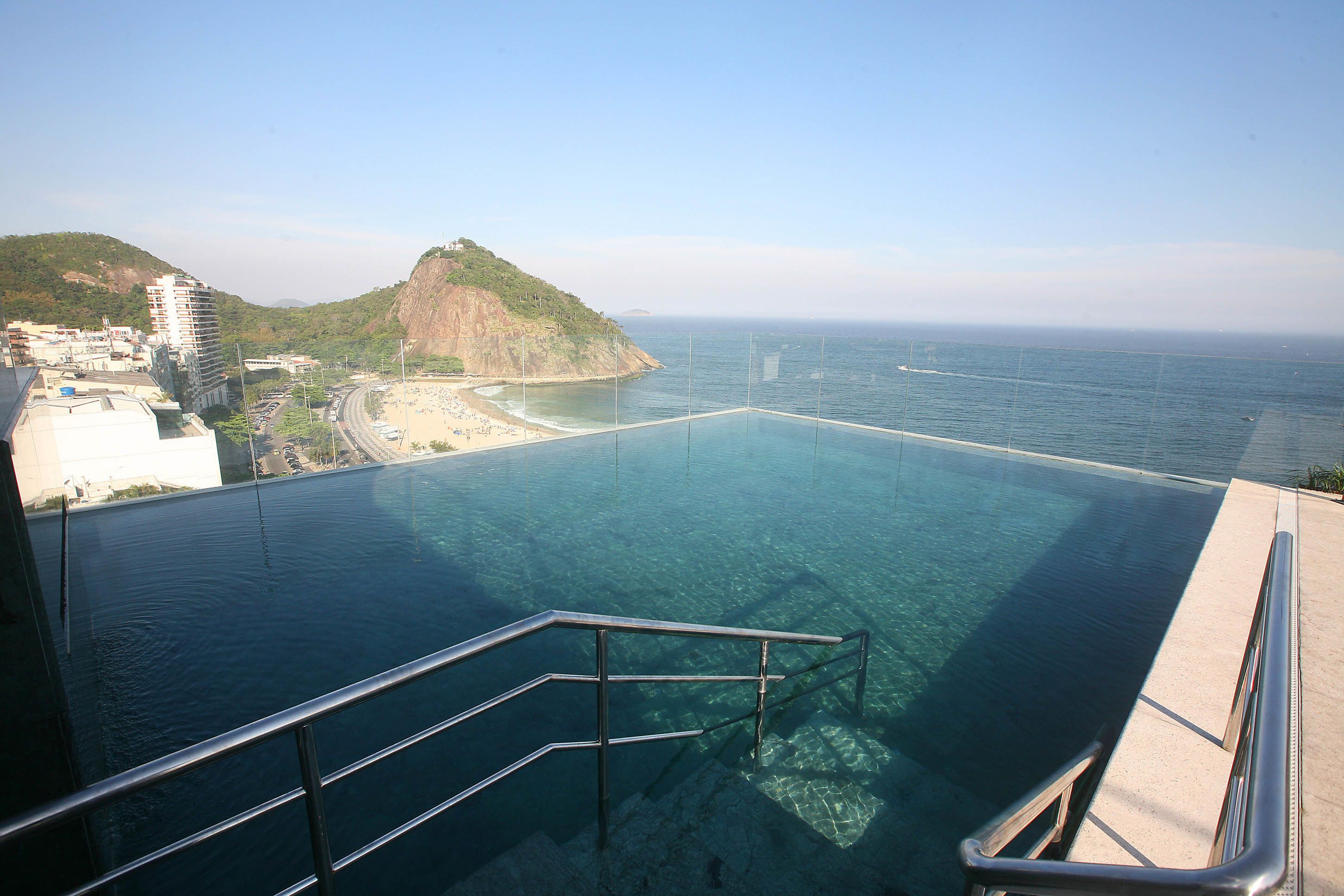 Vista despejada de una piscina con barandilla metálica, que domina una playa y un paisaje montañoso.