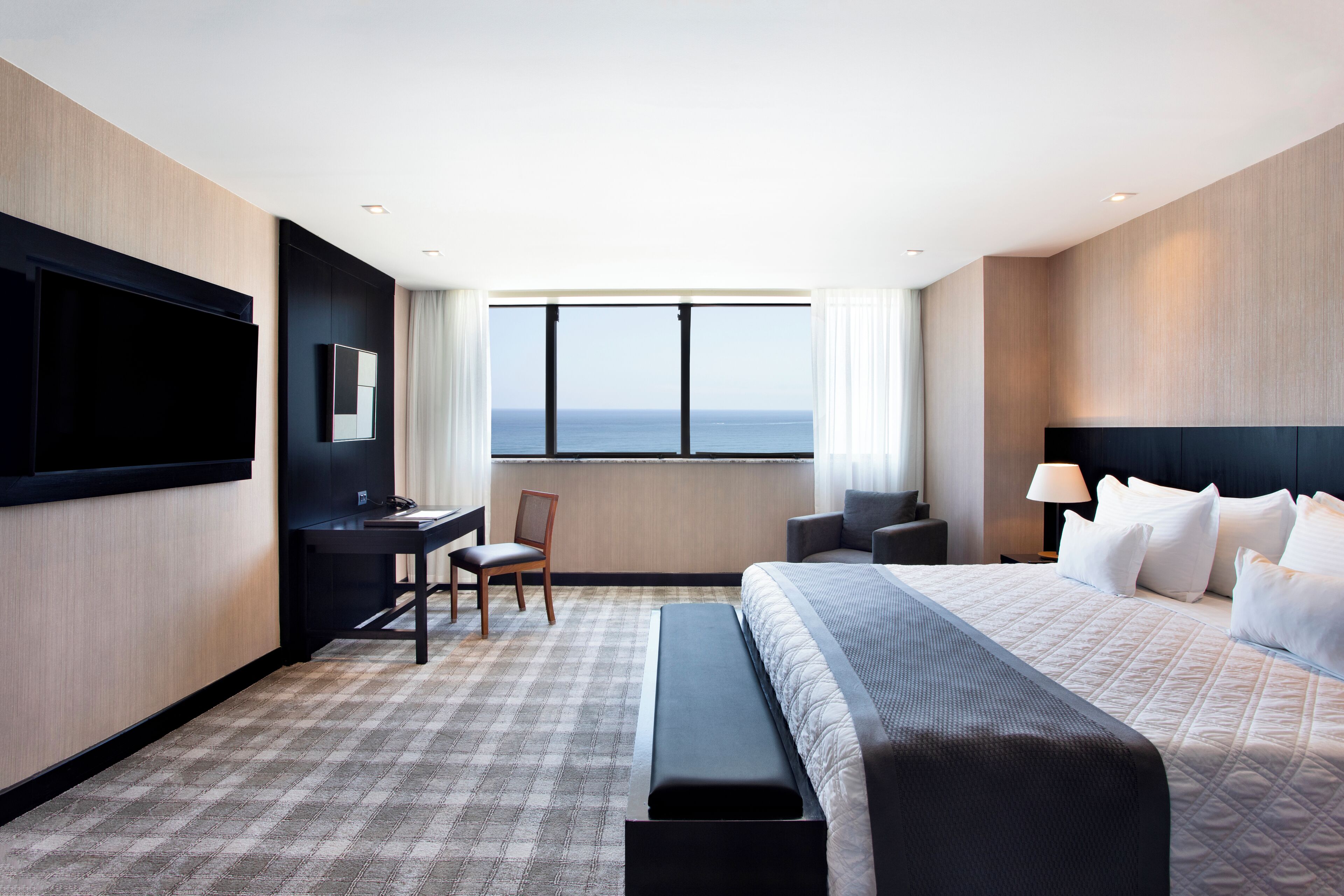 A hotel room with a large bed, a desk, a chair, a TV, and a view of the sea.