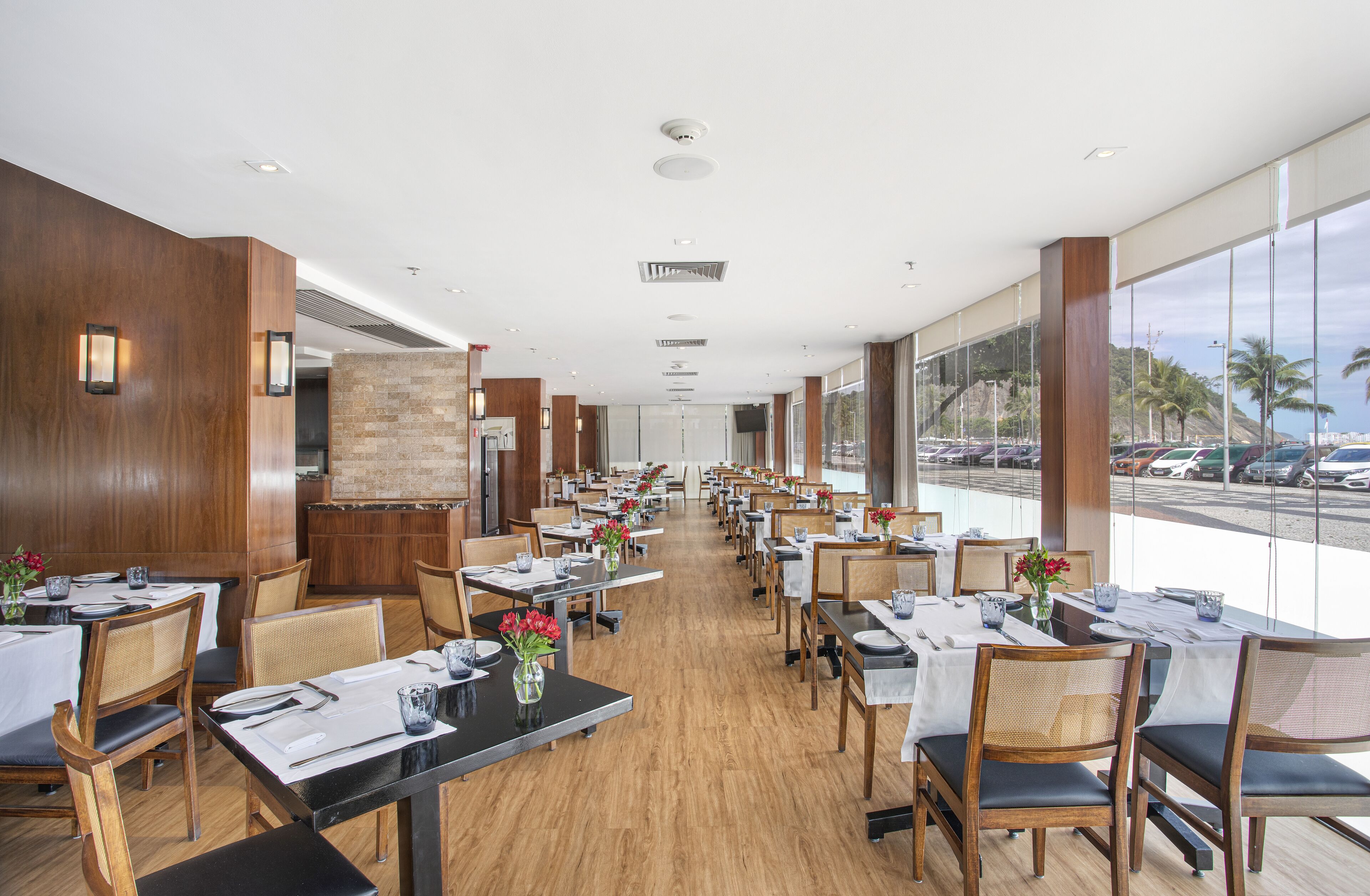 Un restaurante espacioso con paredes de madera y amplios ventanales que ofrecen vistas a coches aparcados y palmeras. Las mesas están ordenadas con manteles blancos y centros de mesa.