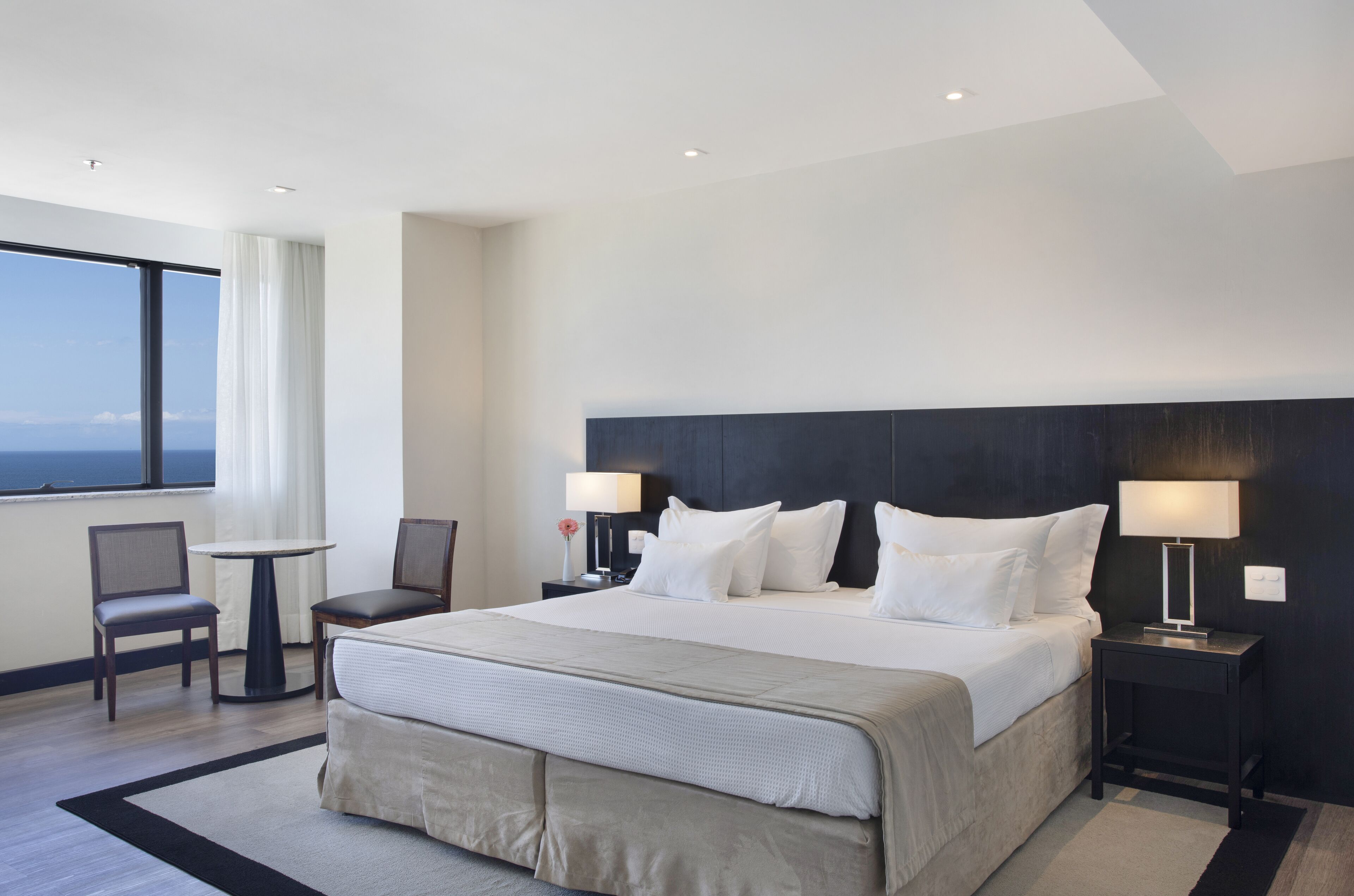 Una moderna camera d'hotel con un letto grande, due comodini con lampade, un piccolo tavolino e una poltrona, e vista sull'oceano da una grande finestra.