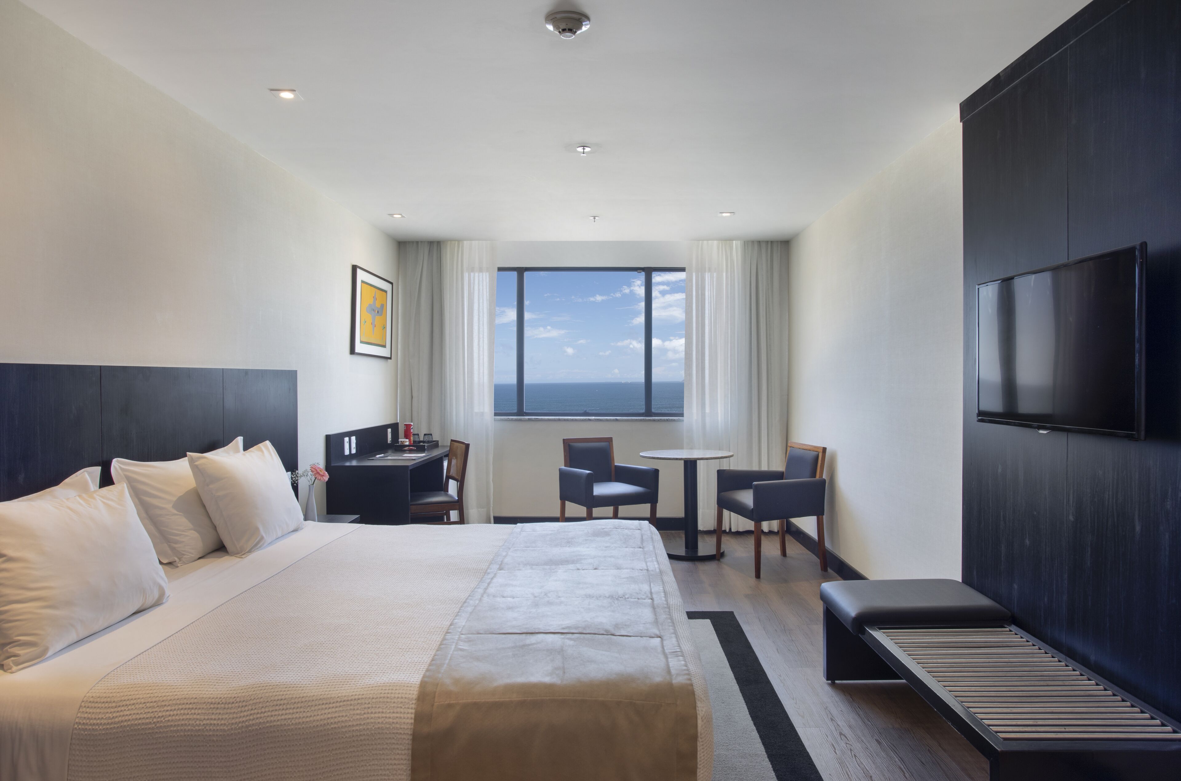 Quarto de hotel com uma cama grande, uma escrivaninha, duas cadeiras, uma televisão e uma janela com vista para o céu.