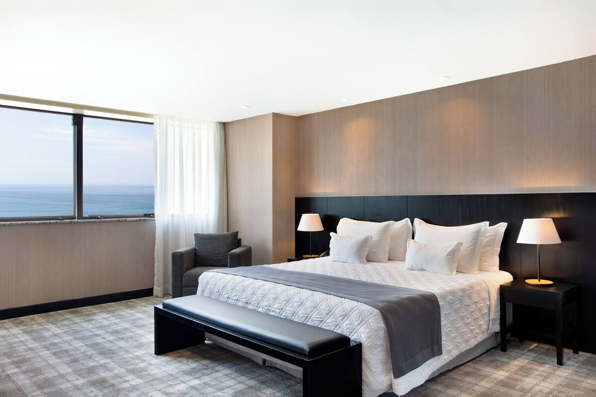 Habitación de hotel moderna con una cama grande, un banco, dos mesitas de noche con lámparas, una silla y vista al océano.