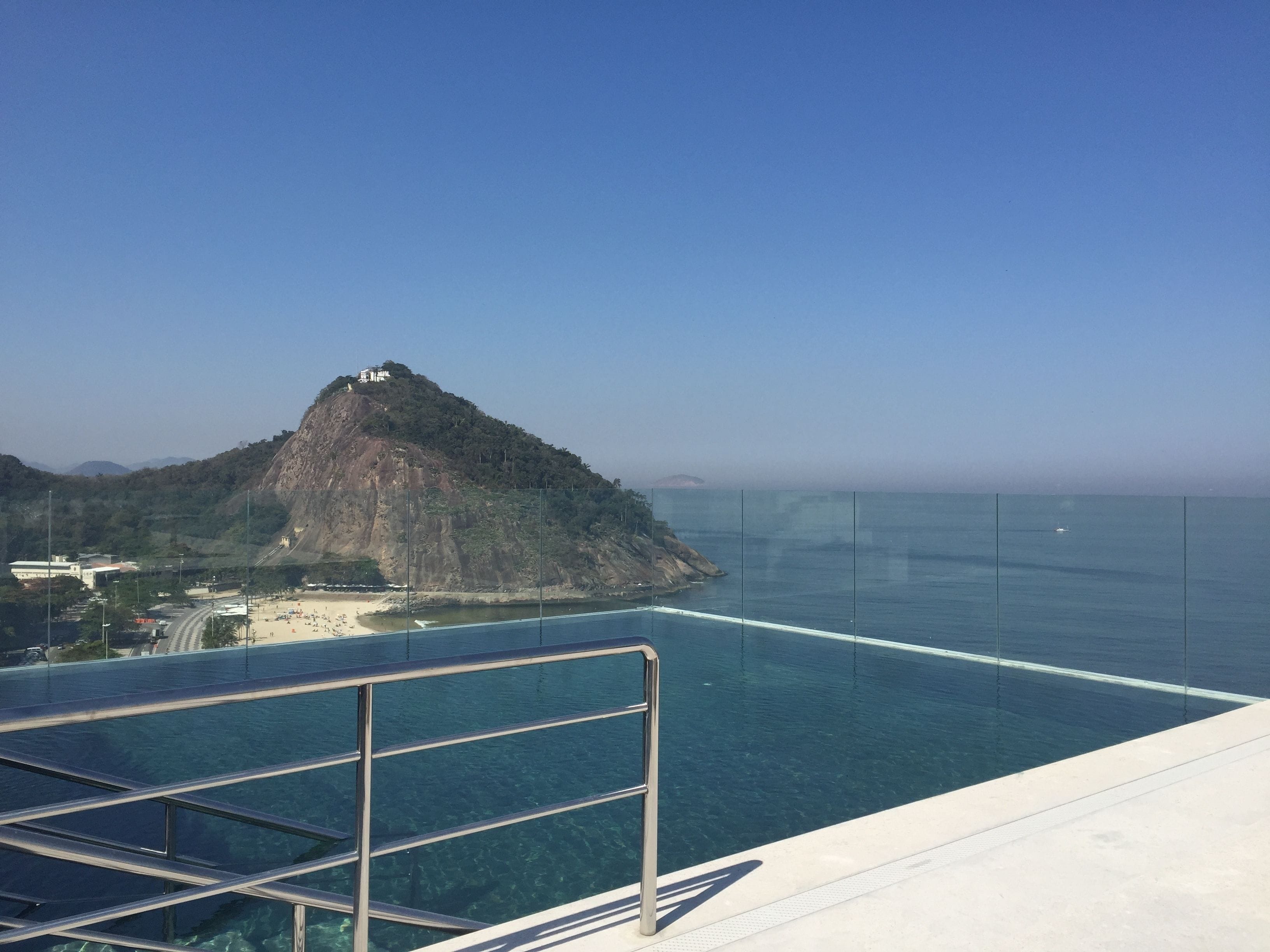 Visão nítida de uma piscina com grade de metal, praia e montanha ao fundo.