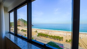 Vista desde la ventana de un hotel sobre una playa con palmeras, una carretera y una montaña.