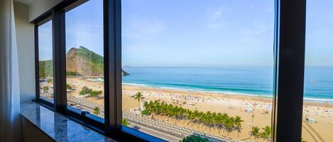 A view from a hotel room window overlooking a beach with palm trees, a road, and a mountain.
