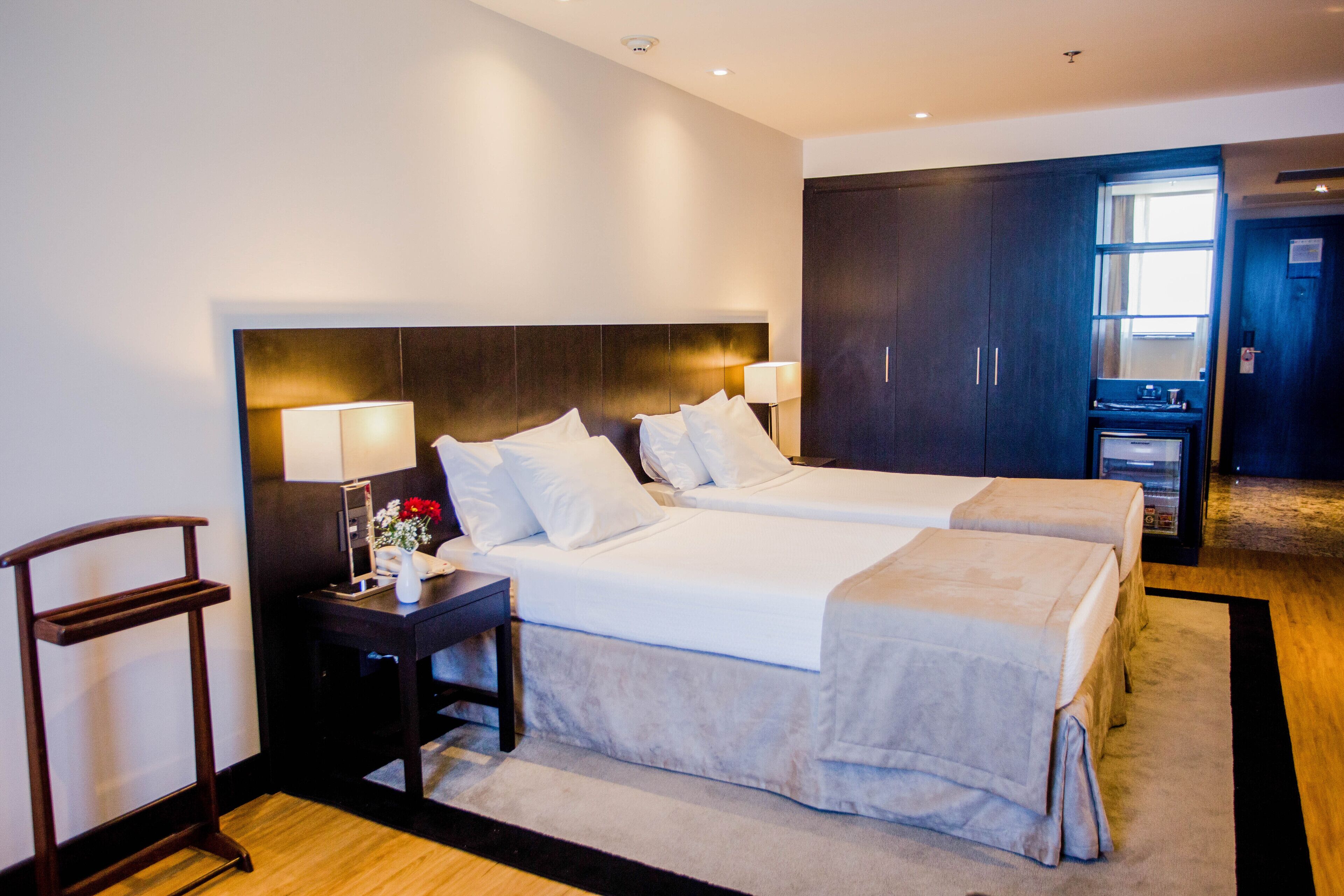 Quarto de hotel com duas camas, mesa de cabeceira com abajur, escrivaninha e guarda-roupa.