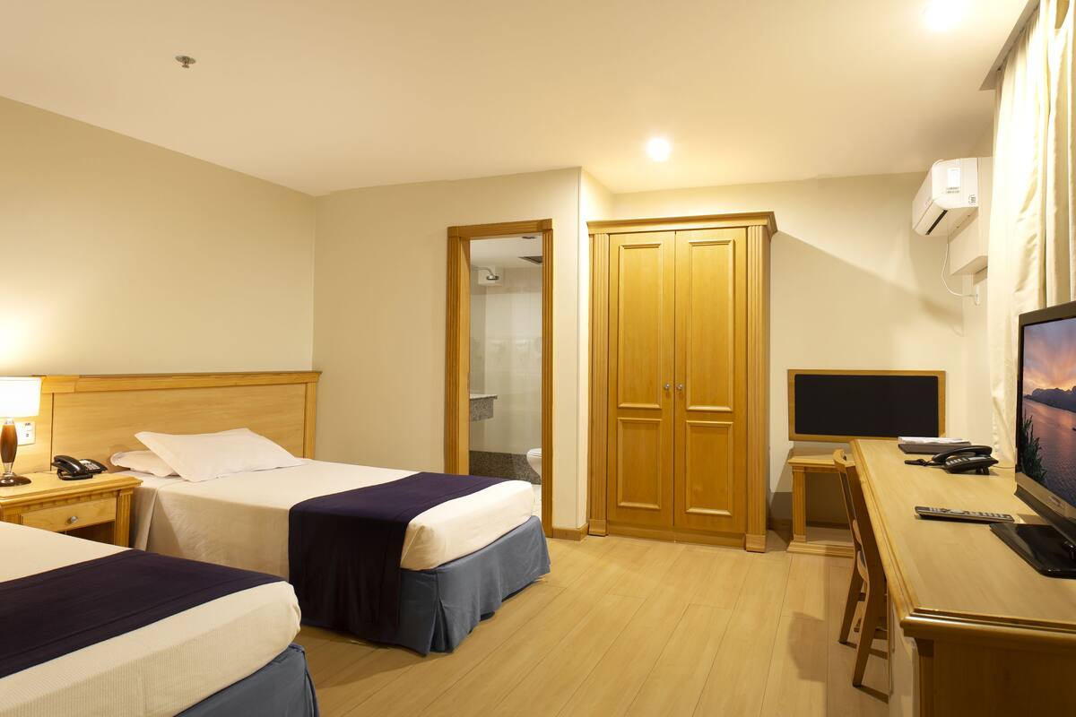 A hotel room with two beds, a desk with a computer, a TV, and a bathroom with a shower and toilet.