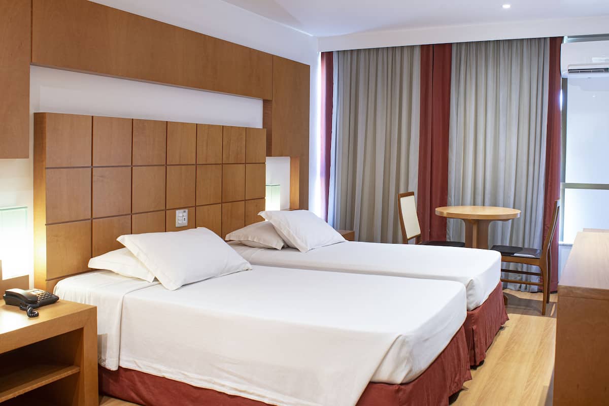 A hotel room with two beds, a desk, a chair, a small table, and a window with curtains.