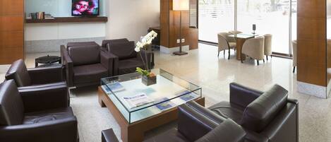 Un moderno hall de hotel con sofás de cuero, una mesa de centro de vidrio, un televisor en un estante y un mostrador de recepción.