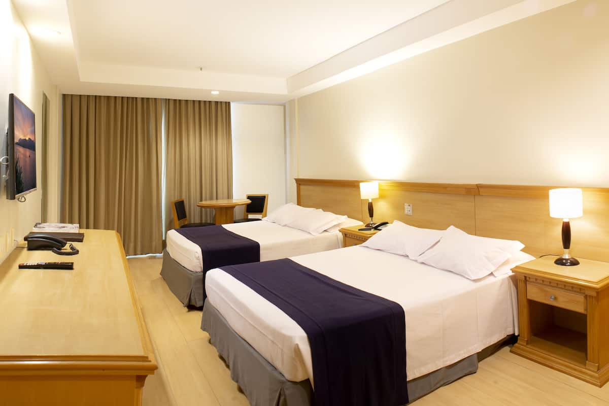 A hotel room with two beds, a desk, a chair, a TV, and a window with curtains.