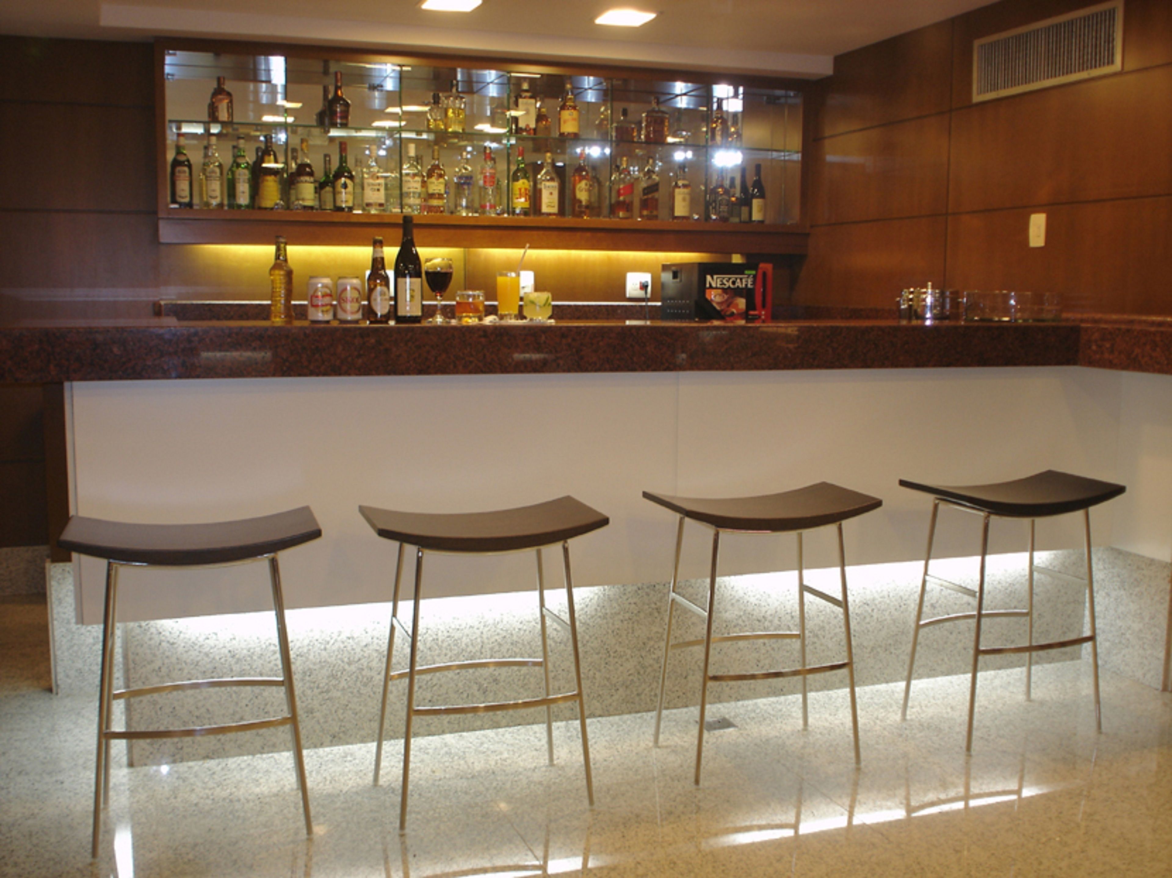Eine Bar mit einer Theke, auf der verschiedene Flaschen und eine Nescafé-Maschine ausgestellt sind. Vor der Theke stehen drei Barhocker.