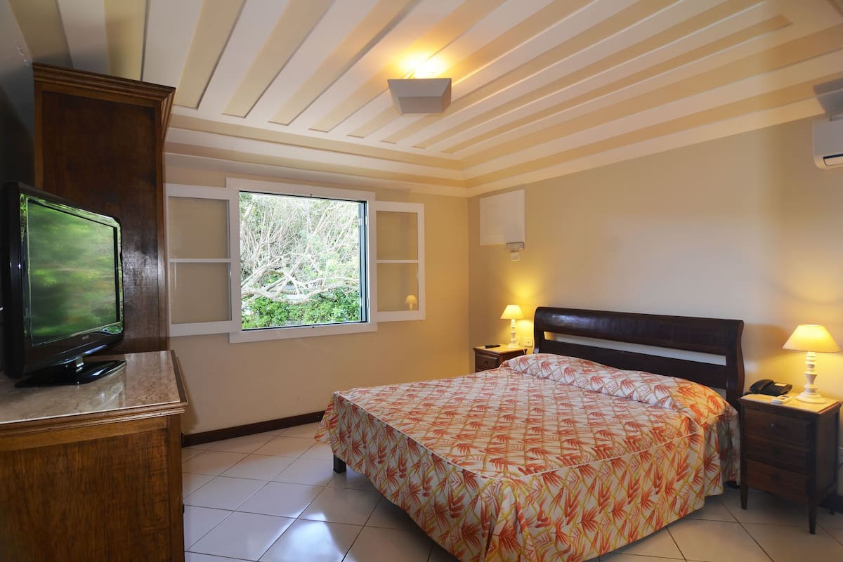 A bedroom with a bed, a TV, a window, and a nightstand with a lamp.