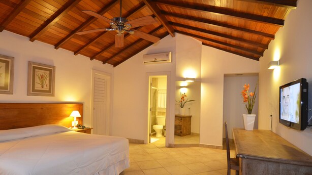 A bedroom with a bed, bedside lamp, wooden ceiling, and a TV on the wall. - Pousada dos Buzios (Búzios)