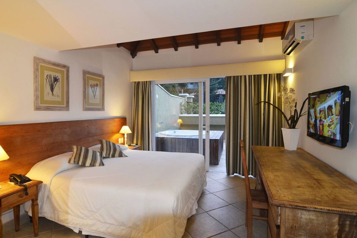 A hotel room with a bed, bedside table, lamp, TV, and a view of a pool and garden.