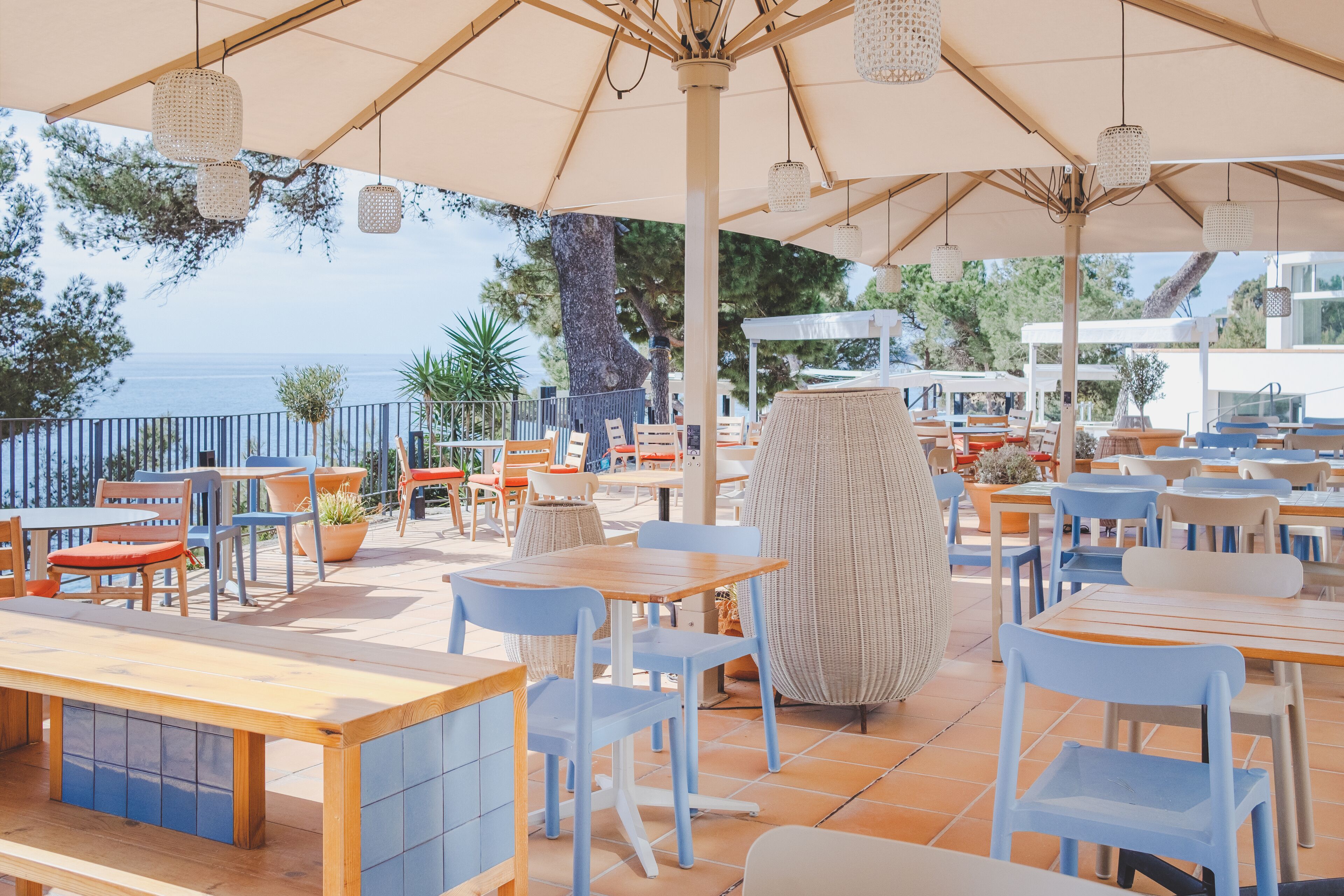 Un área de comedor al aire libre con mesas y sillas, un paraguas grande y vista al mar.