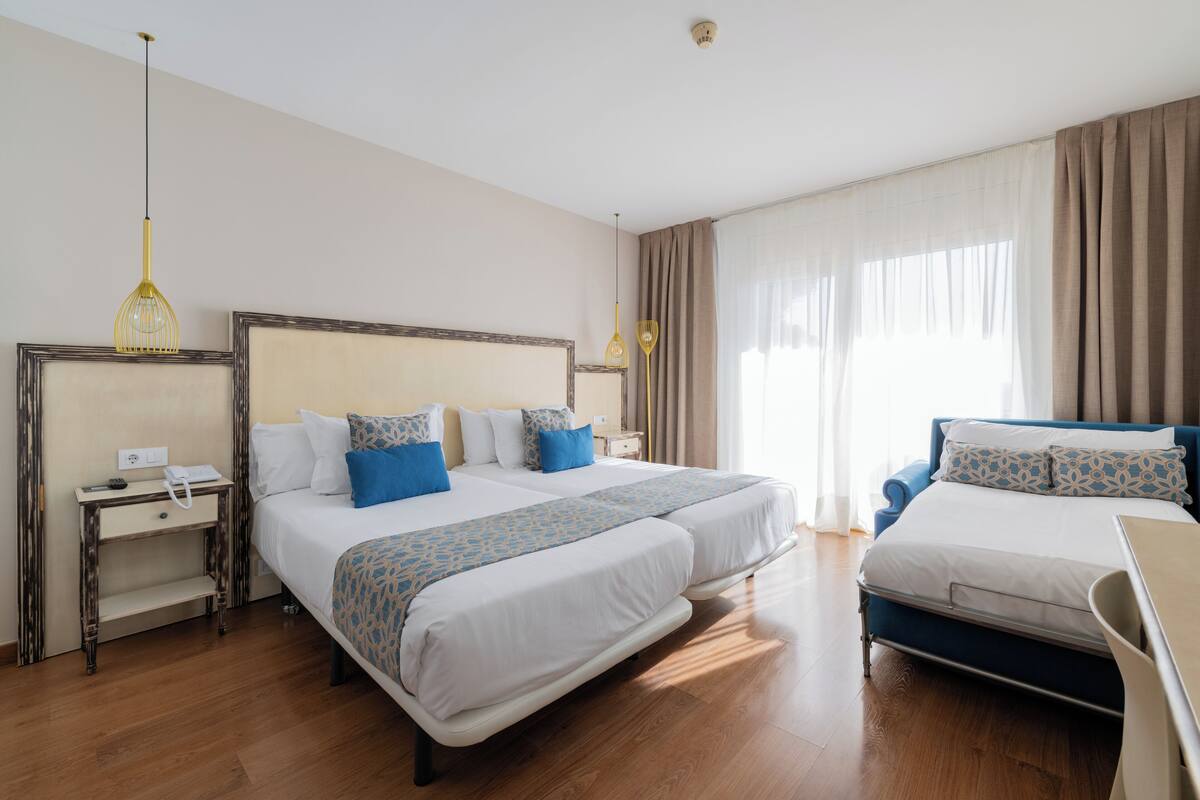 A hotel room with two beds, a bedside table, a lamp, and a window with curtains.