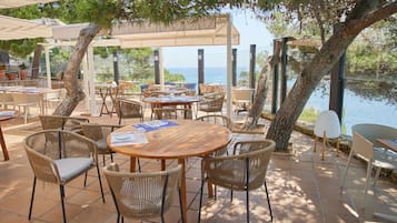 Een terras buiten met ronde houten tafels en rieten stoelen, met uitzicht op een waterpartij.