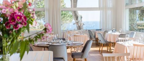 A restaurant with round tables, chairs, and a view of the sea.