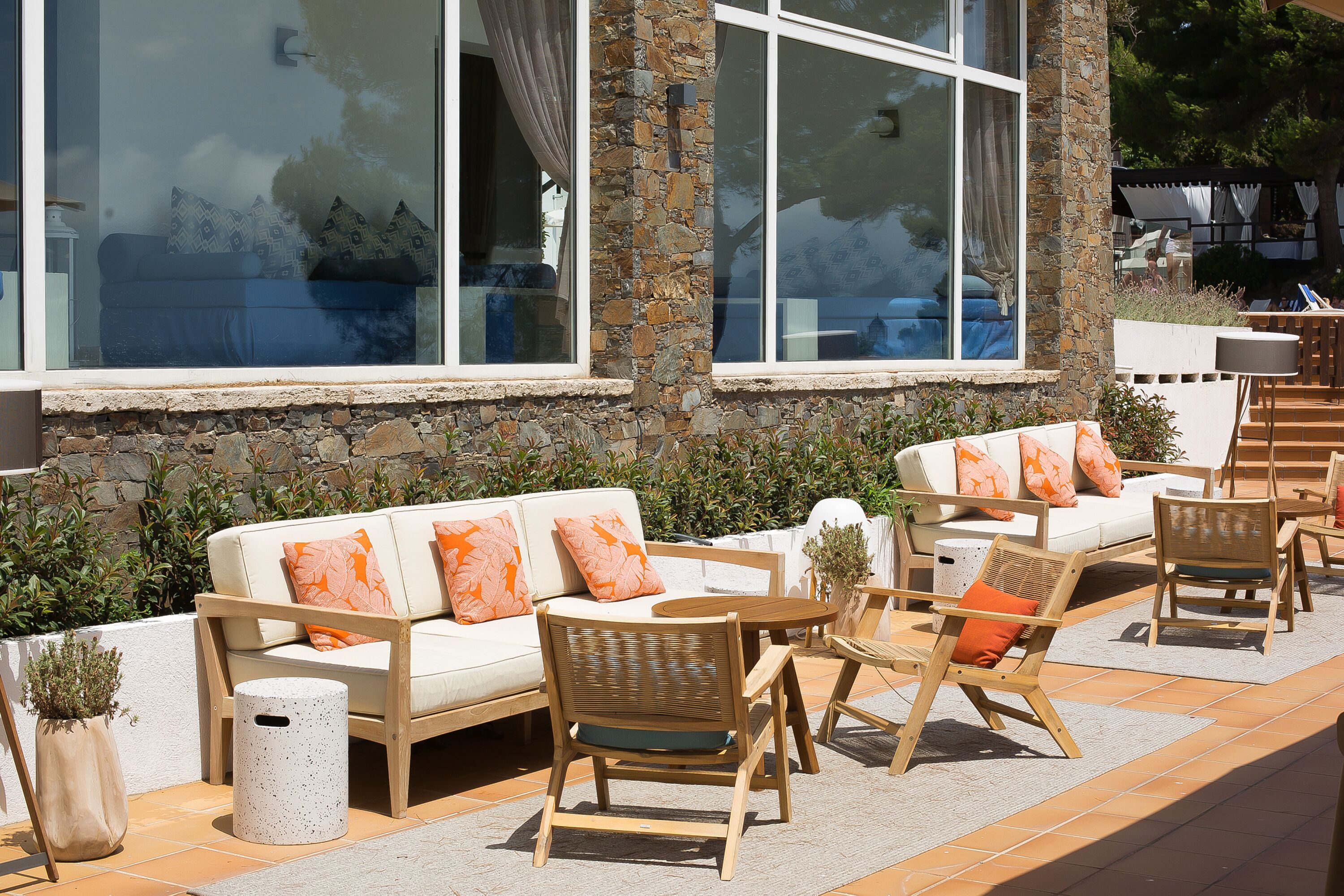 An outdoor seating area with a stone wall, a white sofa with orange cushions, and wooden chairs.
