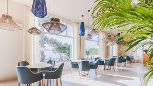 A spacious dining area with round tables and blue chairs, large windows with natural light, and hanging wicker lamps. - Park Hotel San Jorge & Spa by Escampa Hotels (Calonge)