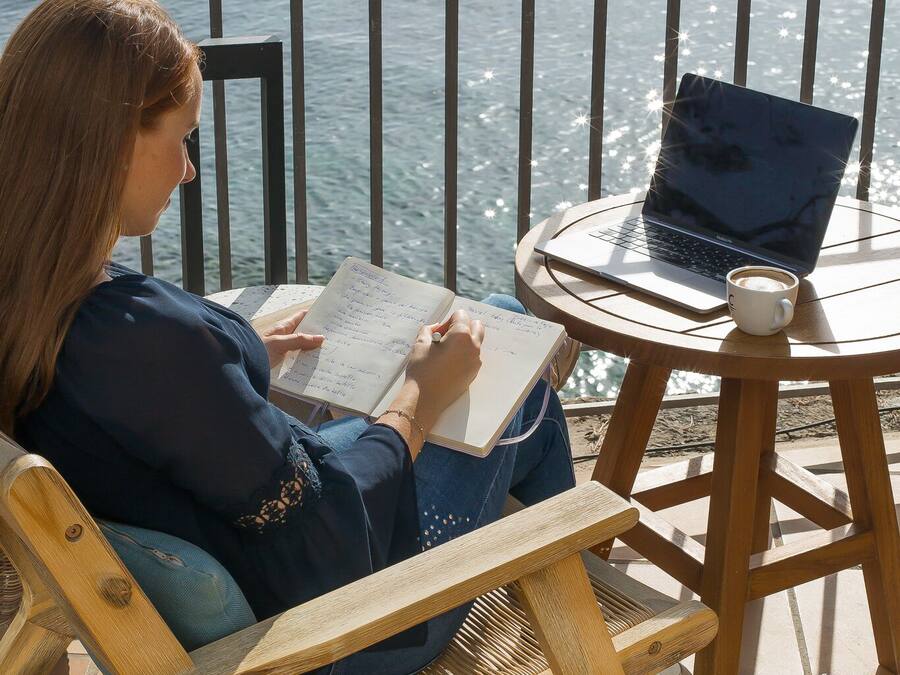 A person seated at a wooden table with a laptop and a notebook, overlooking a body of water.
