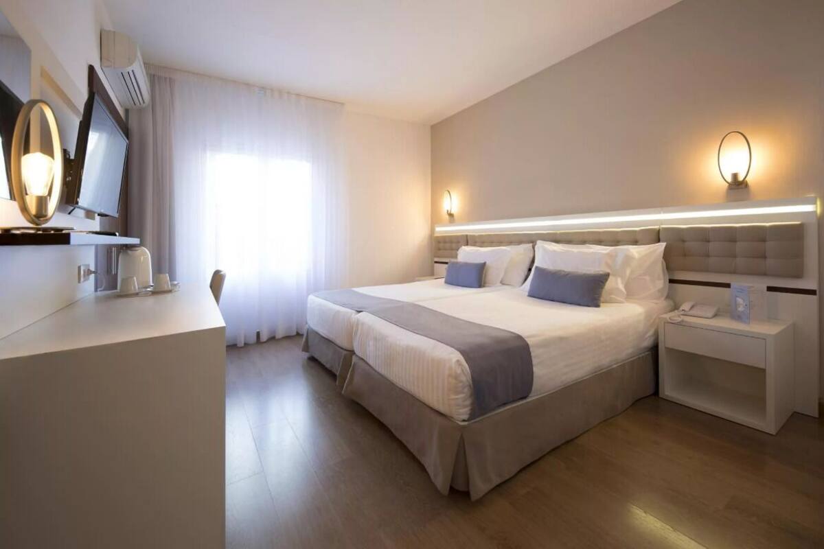 A modern hotel room with a large bed, bedside tables, a flat-screen TV, and a large window with curtains.