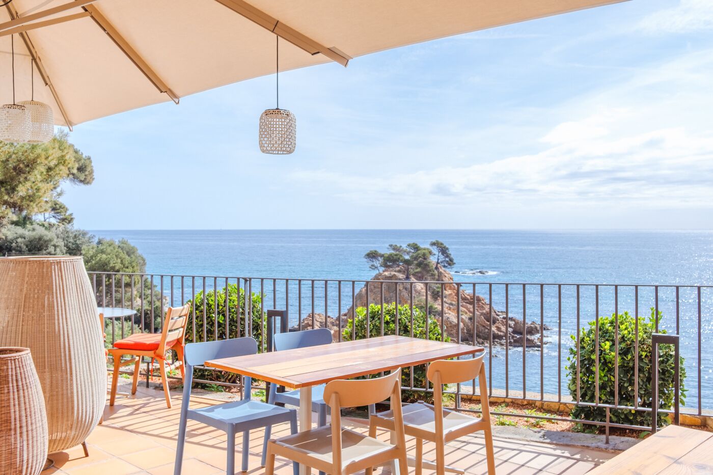Una terraza con mesa y sillas de madera, con vista al mar, un promontorio rocoso y árboles.