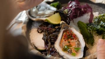 Una persona sosteniendo una ostra con cobertura de caviar rojo, rodeada de diversos mariscos y vegetales en un plato.