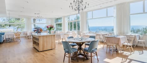 A spacious dining area with round tables and chairs, large windows offering a view of the sea, and a central island with a vase of flowers.