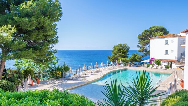A pool area with lounge chairs and umbrellas, surrounded by greenery and a building with a tiled roof. - Park Hotel San Jorge & Spa by Escampa Hotels (Calonge)