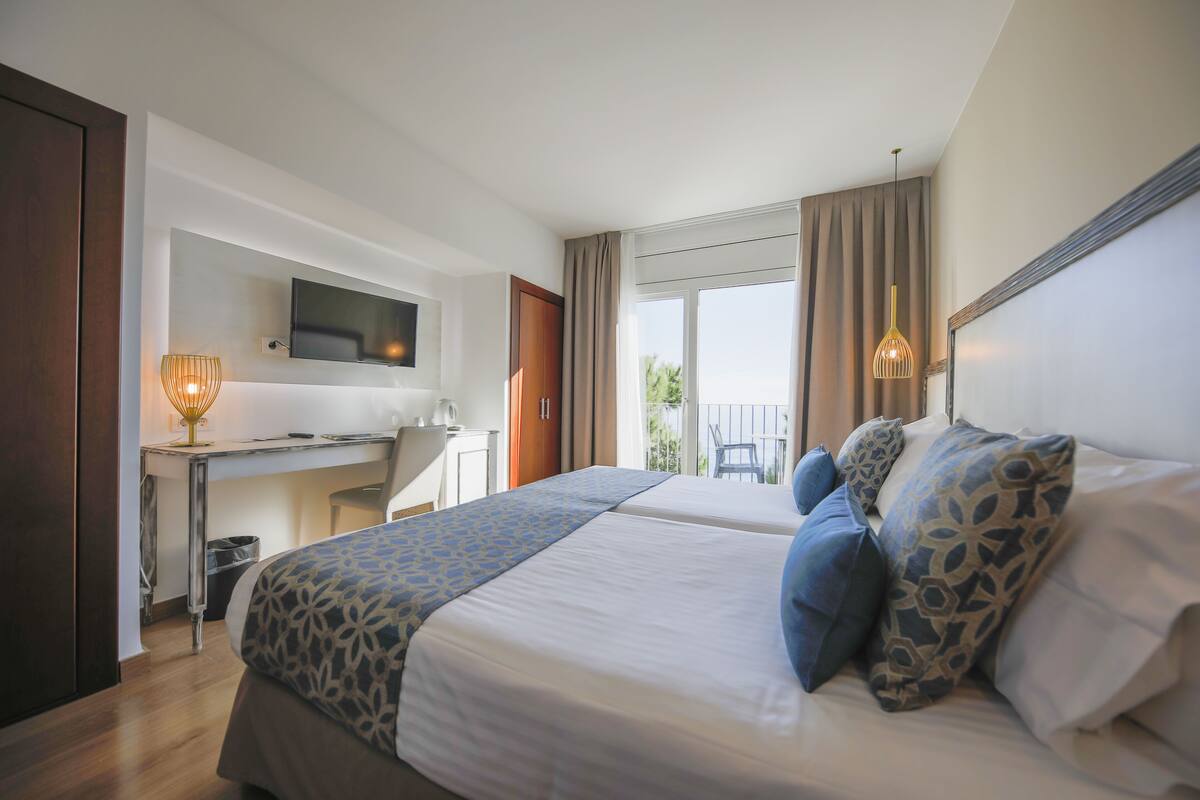 A hotel room with two beds, a desk, a television, and a balcony with a chair.