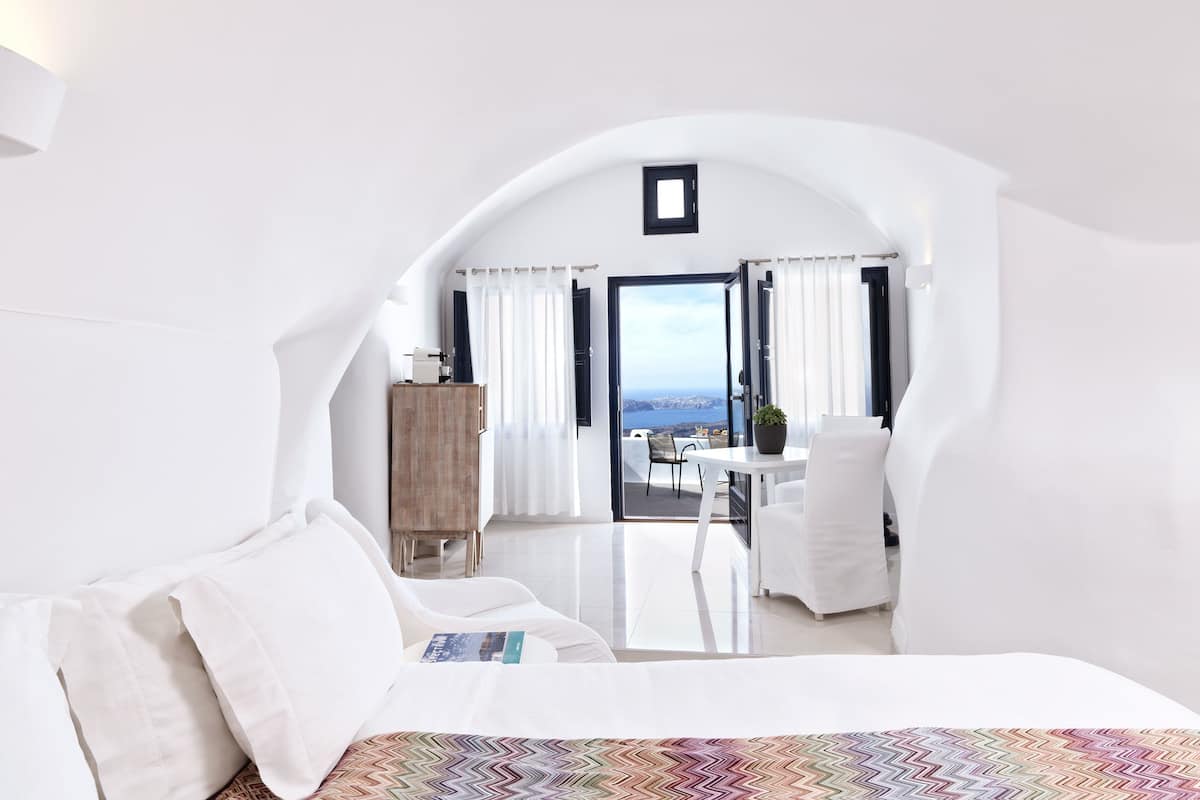 Ein modernes Schlafzimmer in Weiß mit einem großen Bett, einem kleinen Tisch und Meerblick durch eine offene Tür.